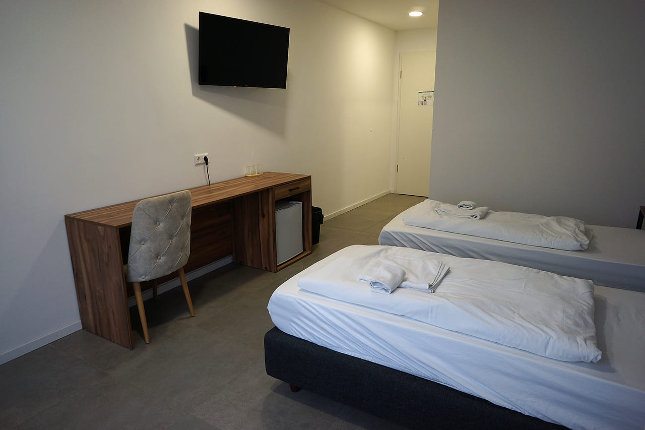Langer Schreibtisch mit Stuhl im Deluxe-Zimmer, gegenüber das Bett, darüber ein Wandfernseher.