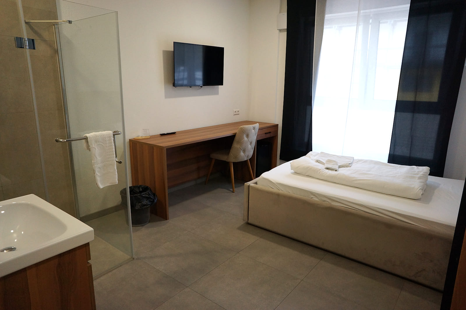 Einzelzimmer mit langem Schreibtisch, Wandfernseher, Bett am Fenster und bodengleicher Dusche hinter Glas.
